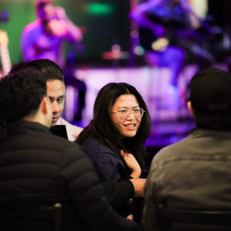 A woman in glasses smiles at a table with friends, with a blurred stage in the background.
