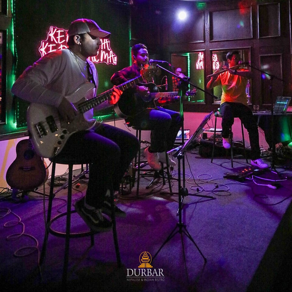 Three musicians performing live on stage with neon lights in a dark room at Durbar Bistro.