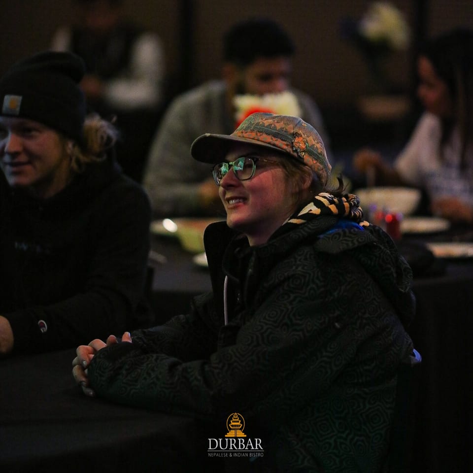 A smiling person in glasses and a patterned hat sits at a table in a dimly lit Durbar restaurant.