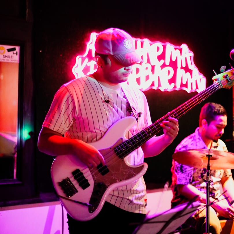 A musician in a baseball cap plays bass guitar on stage with a drummer in the background.