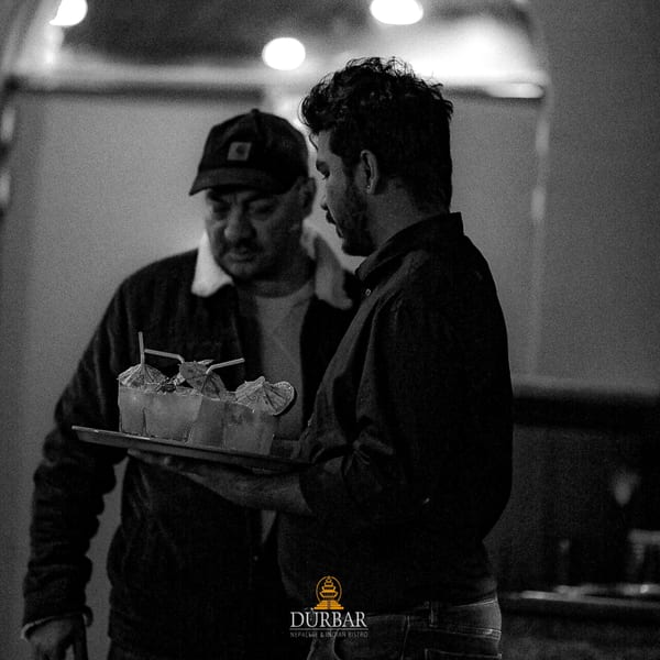 Two men, one carrying a tray of cocktails with umbrellas, in a dimly lit restaurant.