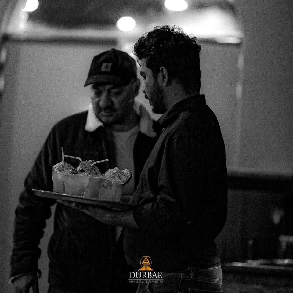 Two men, one carrying a tray of cocktails with umbrellas, in a dimly lit restaurant.