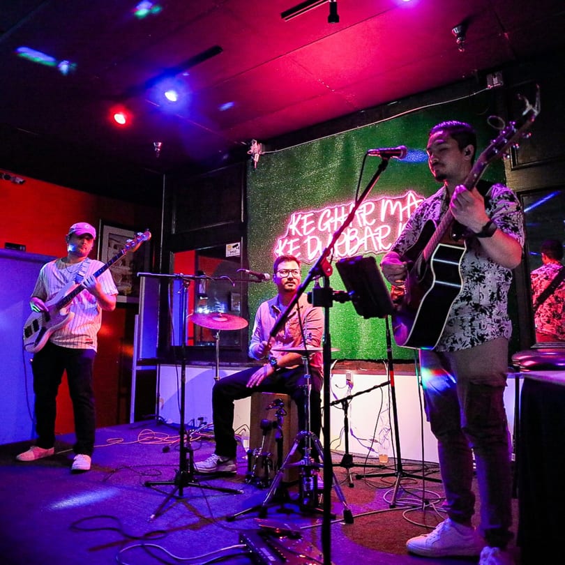 Three musicians performing live on a stage with colorful lighting and a neon sign.